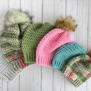 Op de afbeelding: Vijf gebreide mutsen met nep bont pompoenen in verschillende kleuren, waaronder groen, roze en blauw. De mutsen zijn in een rij op een wit houten oppervlak gerangschikt.