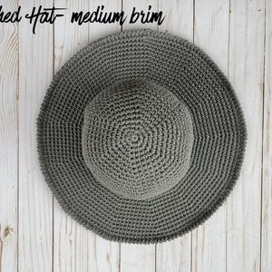 May include: A gray crocheted sun hat with a medium brim. The hat is lying flat on a white wooden surface.