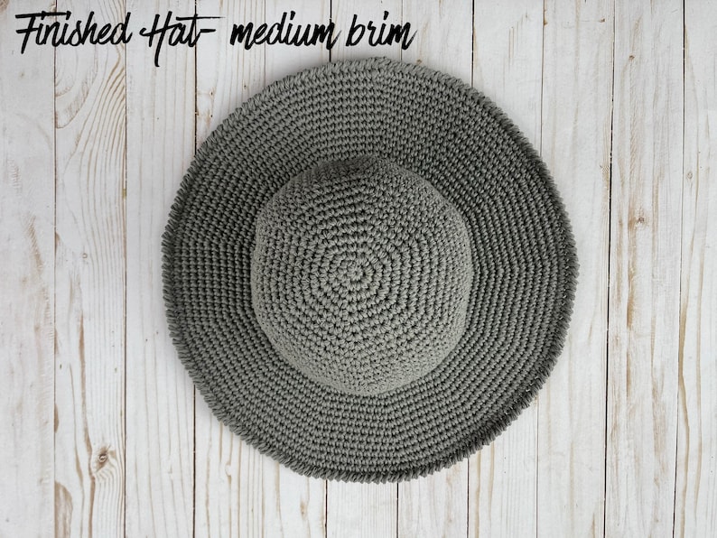 May include: A gray crocheted sun hat with a medium brim. The hat is lying flat on a white wooden surface.