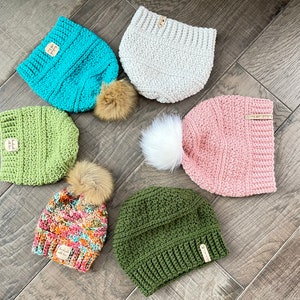 May include: A collection of handmade crochet beanies in various colors, including teal, green, pink, and olive. Each beanie features a faux fur pom-pom and a small wooden tag. The beanies are arranged on a wooden surface.