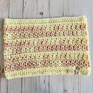 May include: A crocheted neck warmer with alternating stripes of pale yellow and peach yarn. The warmer has a small brown tag attached to the bottom right corner.