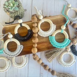 Puede incluir: Un conjunto de juguetes de madera en forma de anillo con ribete de macramé en negro, blanco y turquesa. Los anillos cuelgan de cuentas de madera en una cuerda.