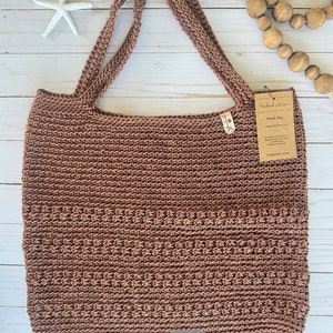 May include: A brown knitted beach bag with long straps. The bag has a textured pattern and a pocket on the front. The words "Seaside Beach Bag" are at the bottom of the image. A tag is attached to the bag.