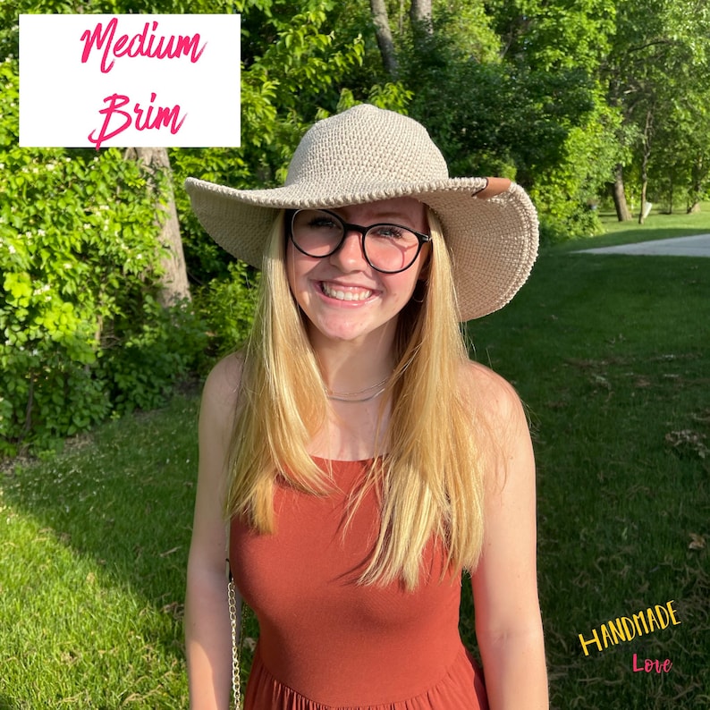May include: A woman wearing a beige crochet sun hat with a medium brim, a rust-colored sleeveless dress, and glasses. The hat has a brown leather band. The background is a green grassy area with trees.