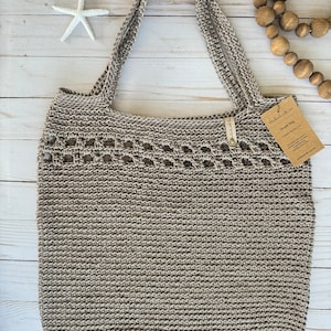 Pode incluir: Uma bolsa de praia de crochê bege com uma etiqueta de couro marrom que diz "Handmade with love" e "Beach Bag".