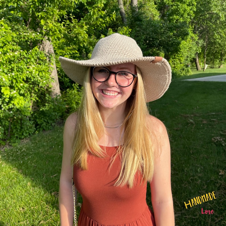 May include: A woman wearing a beige crochet sun hat with a brown leather accent, a rust-colored sleeveless dress, and glasses. The hat has a wide brim and is crocheted with a delicate pattern. The woman is smiling and looking at the camera. The background is a green grassy area with trees in the distance. The text "HANDMADE Love" is visible in the lower right corner of the image.