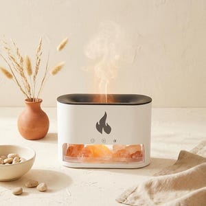 May include: A white rectangular essential oil diffuser with a black top rim and a flame icon on the front. The base features a transparent window displaying several orange Himalayan salt rocks, while a mist rises from the top center opening.