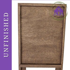 May include: A rectangular, unfinished wooden sign with a smooth, light brown surface and a raised border frame. The sign features a small horizontal ledge at the bottom and includes the text DIY Paint Kit and UNFINISHED on the purple border.