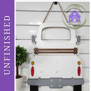 May include: An unfinished wooden DIY paint kit shaped like the rear view of a vintage pickup truck. The cutout features a white body, a grey bumper, black tyres, brown tailgate slats, and red and yellow taillights, suspended by a jute twine hanger.