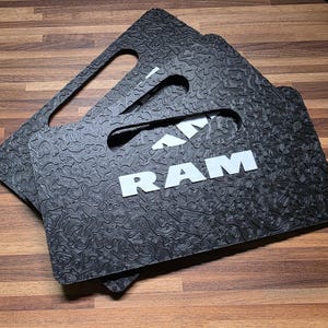 May include: Two rectangular black floor mats featuring a textured topographic map pattern and a white RAM logo. Each mat includes a cutout handle at the top edge. The surface of the mats has a raised, irregular, and wavy line design throughout.