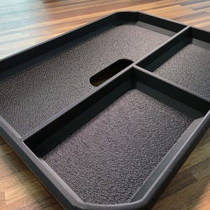 May include: A rectangular black plastic tray featuring three distinct compartments with a textured surface finish. The tray includes a central integrated handle cutout, raised edges, and a structured design for organizing items within the sections.