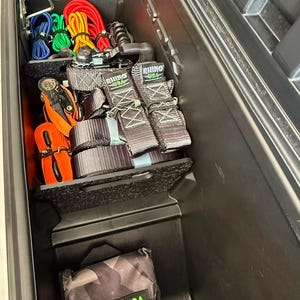 May include: A black plastic truck bed storage compartment containing several Rhino USA ratchet straps with grey webbing, orange straps, and colorful bungee cords. A camouflage patterned storage bag with a Rhino USA label sits below.
