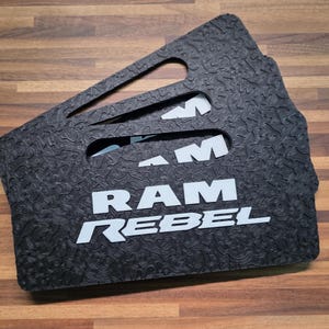 May include: A set of black rectangular automotive floor mats featuring a textured, topographic-style surface pattern. The mats display the white text RAM REBEL in a bold, sans-serif font and include integrated handle cutouts at the top edge.