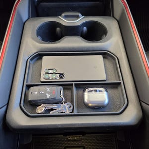 May include: A black plastic center console organizer tray featuring three distinct compartments. The top section holds a rectangular smartphone, while the two lower sections contain a black vehicle key fob with metal keys and a silver wireless earbud case.