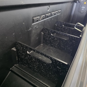 Könnte beinhalten: Ein schwarzes Kunststoff-Staufach für Ladeflächen mit der Aufschrift RAMBOX und mehreren herausnehmbaren Trennwänden. Die Trennwände haben eine strukturierte, schwarz-weiß gesprenkelte Oberfläche und integrierte Griffaussparungen zur Anpassung.