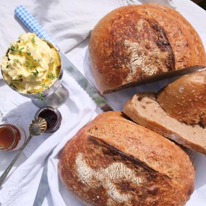May include: Two round loaves of crusty brown sourdough bread, one sliced, alongside a silver bowl filled with yellow butter topped with green herbs, a small jar of dark jam, a small jar of orange jam, and a silver spoon with a blue chequered handle.