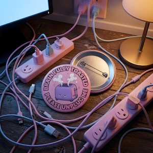 Può includere: Una spilla rotonda rosa con l'illustrazione di un tostapane viola da cui esce vapore e il testo "BRAIN FULLY TOASTED PLEASE UNPLUG ME" in lettere maiuscole nere, mostrata con il meccanismo a spilla in metallo visibile sul retro.