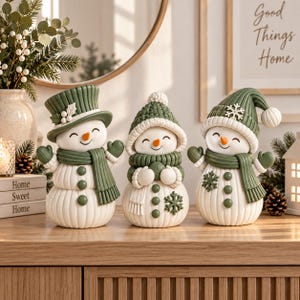 Puede incluir: Tres figuras decorativas de muñecos de nieve con cuerpos blancos de textura acanalada vertical y detalles en verde oscuro. Cada muñeco de nieve lleva una bufanda y un gorro verdes, con narices de zanahoria naranja y botones verdes.