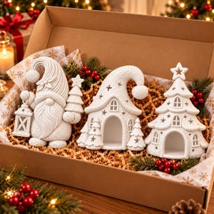 May include: A set of three white ceramic Christmas decorations in a cardboard box, including a gnome holding a lantern and a tree, a small house with a curved roof, and a tiered Christmas tree structure, all nestled in brown paper filler.