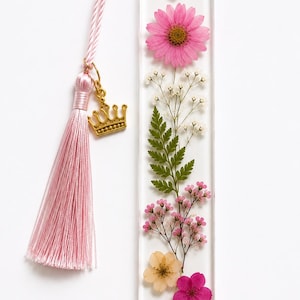 May include: A rectangular clear resin bookmark containing pressed pink daisy, white baby's breath, green fern, and small pink flowers. A light pink cord is attached to the top with a gold-toned metal crown charm and a matching pink tassel.