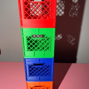 May include: A vertical stack of four miniature plastic crates in bright colors. The top crate is red, followed by a green crate, a blue crate, and an orange crate at the base. Each crate features a rectangular handle cutout and a diamond grid pattern.