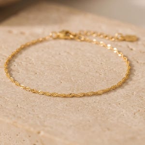 May include: A delicate gold-toned rope chain bracelet featuring a twisted link design. The bracelet includes a thin chain, a small circular clasp, and an adjustable extension chain with a tiny flat tag at the end, showcasing a polished finish.
