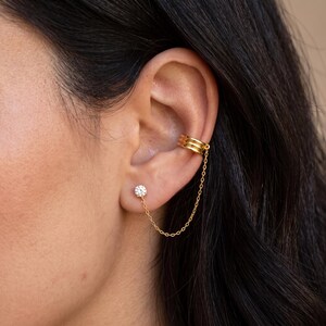 May include: A gold-toned ear cuff featuring three horizontal bands connected by a delicate chain to a single round, clear stud earring. The jewelry piece is worn on the ear, with the cuff positioned on the upper cartilage and the stud on the lobe.