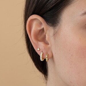May include: A gold ear stack featuring three distinct pieces: a small stud earring with a single white opal, a small hoop earring set with five white opal stones, and a plain polished gold hoop earring, all worn in a single ear lobe.