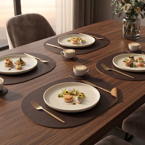 May include: A dining table set with four oval dark brown leather placemats, each featuring a cream-colored ceramic plate with gourmet food, a gold fork, and a small white coffee cup on a matching coaster, arranged for a formal meal service.