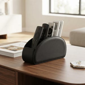 May include: A black semi-circular remote control organizer with a smooth, matte finish. The holder features multiple interior slots containing five various remote controls in black, grey, and white, arranged in a cascading row for easy access.