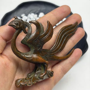 May include: A bronze-toned metal figurine of a mythical bird with an elongated neck, a curved beak, and detailed wings. The creature features textured plumage patterns and stands on a stylized base with swirling decorative elements.
