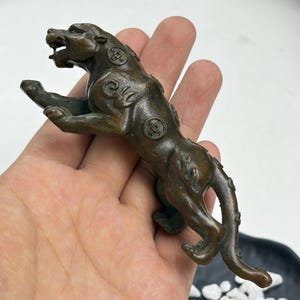 May include: A bronze-toned metal figurine of a leaping panther with an open mouth and bared teeth. The body of the panther is decorated with embossed circular coin-like motifs and swirling patterns along its back and flanks, captured in a dynamic pose.