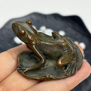 May include: A small, dark bronze-toned metal figurine of a frog resting on a circular lily pad. The frog is depicted in a seated position with its front legs extended forward and its back legs tucked, featuring a textured, weathered surface finish.
