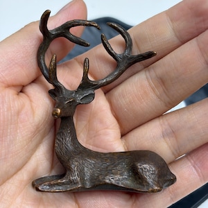 May include: A small, dark bronze-toned metal figurine of a deer in a resting position. The deer features detailed, textured fur and prominent, branching antlers that curve upward. The sculpture has a matte, weathered finish throughout its surface.