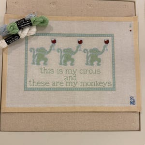 May include: A needlepoint canvas featuring three light green monkeys holding wine glasses, with the text "this is my circus and these are my monkeys" in a light blue border. Three small skeins of silk embroidery thread in blue, green, and white are included.