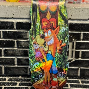 Puede incluir: Una tabla de fingerboard holográfica con un gráfico vibrante de Crash Bandicoot y Tawna de pie en un entorno de selva con una máscara tiki encima. El texto SUGAR FR33 está impreso en las esquinas inferiores de la superficie colorida.