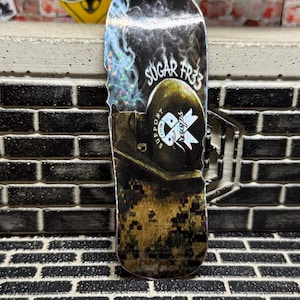 May include: A fingerboard deck featuring a holographic finish with a graphic of a military helmet, the text "SUGAR FR33" and "SUPPORT OUR TROOPS" with a ribbon, and a digital camouflage pattern at the bottom with a small USMC logo printed on the surface.