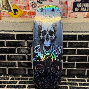 May include: A fingerboard deck featuring a holographic finish with a central graphic of a skull, chains, and roses. The text "Sugar" and "FB33" is printed in a stylised font across the centre of the deck, which has a rounded top and bottom.