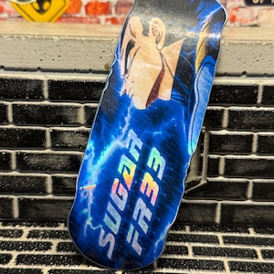 May include: A fingerboard deck featuring a graphic of a character with dark hair in profile against a blue lightning background. The text SUGAR FR33 is printed in a holographic font across the lower portion of the deck, which has a smooth, curved shape.