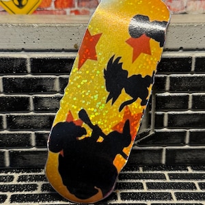 May include: A fingerboard deck featuring a vibrant yellow and orange holographic glitter finish. The surface is decorated with black silhouettes of cartoon characters and several orange star shapes scattered across the background.