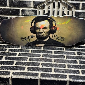 May include: A rectangular decorative plaque featuring a graphic illustration of Abraham Lincoln wearing large over-ear headphones. The text Sugar Fr33 is printed on the image. The surface has a metallic, iridescent finish with a dark, weathered background.