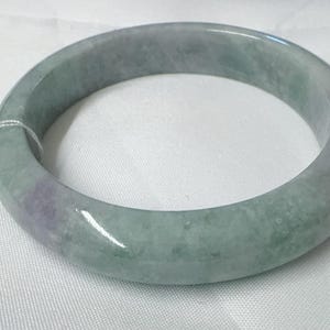 May include: A smooth, polished jade bangle bracelet featuring a rounded profile with mottled light green and subtle lavender color variations. A small, rectangular white paper tag with handwritten text is attached by a thin white string.