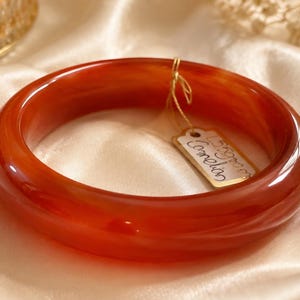 Puede incluir: Un brazalete rígido de cornalina de color rojo anaranjado intenso, pulido y liso, con un perfil redondeado. Una pequeña etiqueta de papel rectangular de color crema está unida al brazalete con un fino alambre dorado, con la palabra Carnelian escrita a mano.