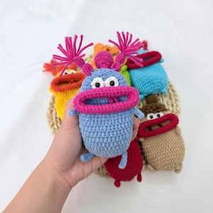 May include: A collection of small, crocheted monster-like figures in various colors including light blue, yellow, red, brown, and turquoise. Each figure features a wide, prominent mouth, two black bead eyes, and two tufted antennae on top.