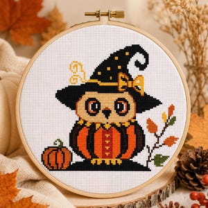 May include: A cross-stitch pattern in a wooden embroidery hoop featuring a stylised owl wearing a black witch hat with a yellow bow, sitting inside a pumpkin. The design includes a small pumpkin to the left and a branch with autumn leaves.