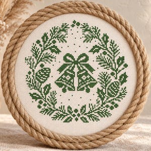 May include: A circular cross-stitch embroidery piece featuring a green wreath design with two central bells and pinecones, framed by a thick, twisted natural jute rope border on a white fabric background.