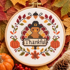 May include: A circular wooden embroidery hoop containing a cross-stitch pattern of a turkey wearing a hat, surrounded by a wreath of autumn leaves, acorns, and pumpkins. The turkey holds a sign with the text Thankful in black lettering.
