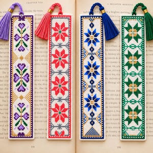 May include: Four rectangular cross-stitch bookmarks featuring geometric patterns in purple, red, blue, and green. Each bookmark is finished with a matching coloured tassel attached to the top with a gold-toned metal bead, resting on an open book.