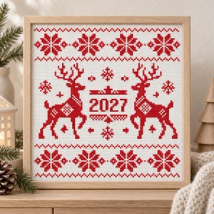 May include: A square framed cross-stitch embroidery featuring a red and white pattern. The design includes two reindeer facing each other, snowflakes, and the year 2027 centered between them. The embroidery is encased in a light-colored wooden frame.