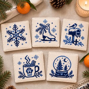 May include: A set of five square fabric coasters featuring blue cross-stitch embroidery on a cream background. The designs include a snowflake, an ice skate, a mailbox with a bird, a steaming mug, and a snow globe, all with winter motifs.